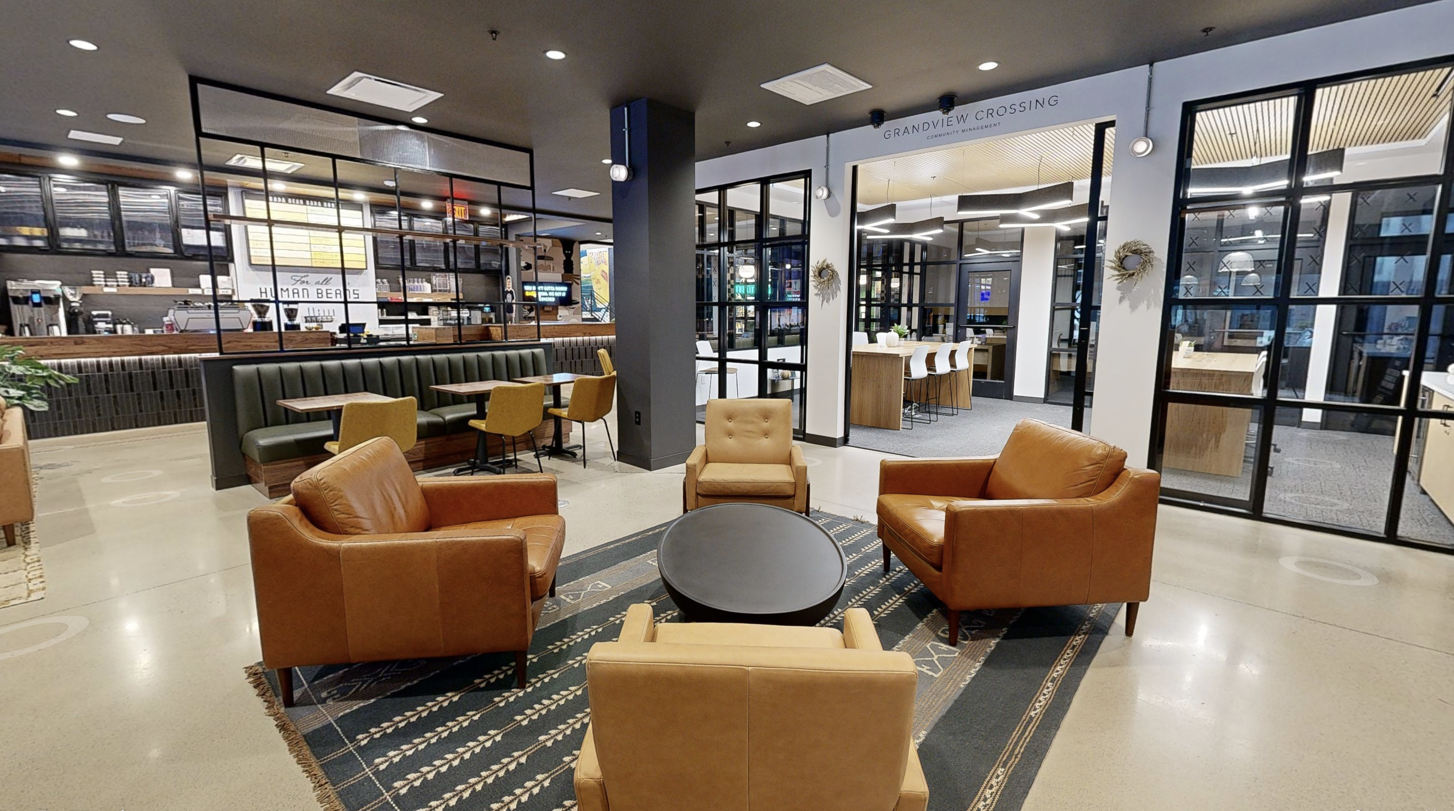 Modern lounge area in Columbus apartments features four brown chairs around a round table. Glass walls elegantly separate the space from a coffee shop counter. The minimalist decor, with its dark ceilings and light flooring, creates an inviting atmosphere for residents. Management office at Grandview Crossing by Thrive companies inside Bada bean bada booze